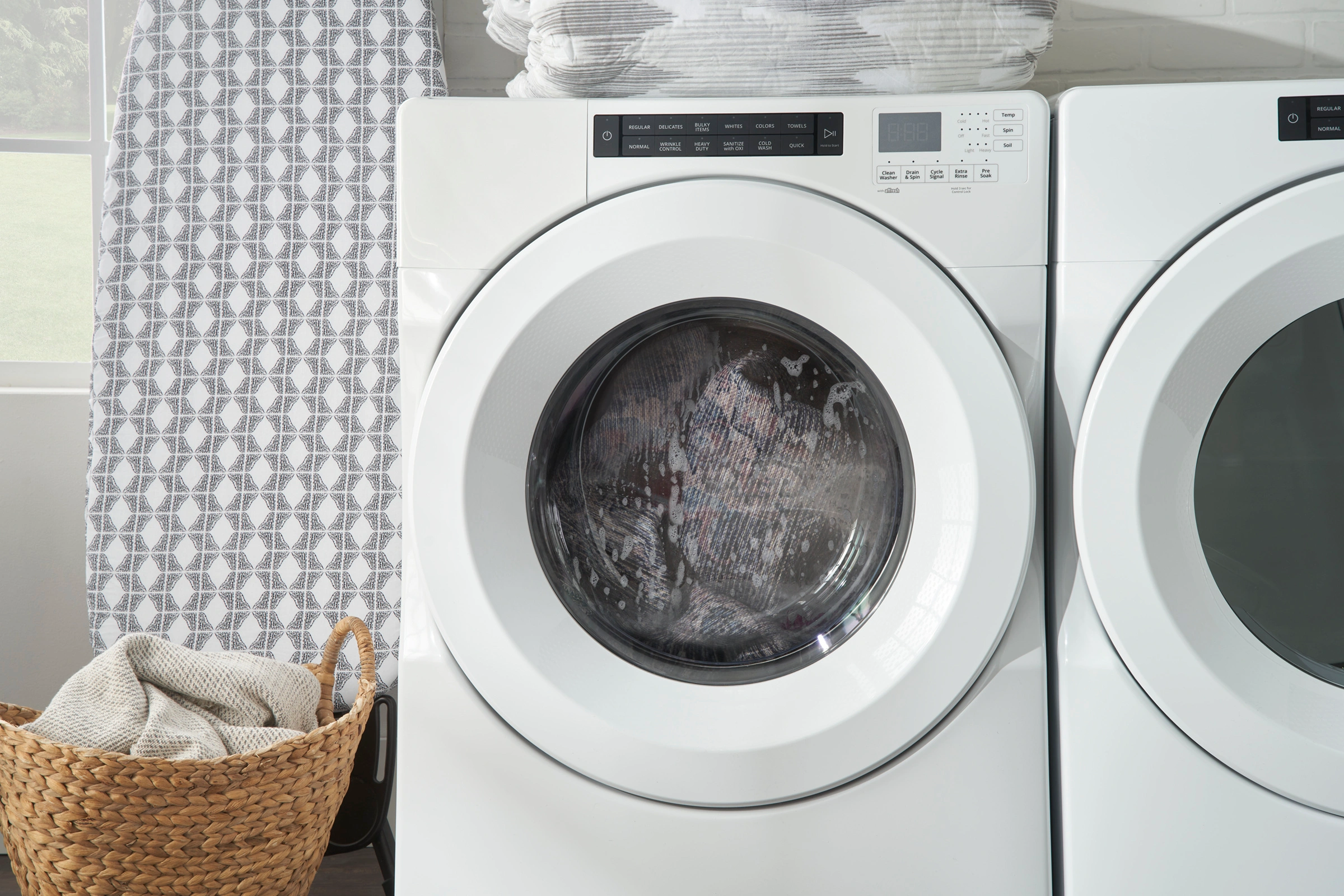 Nourison washable rug tumbling inside a front-load washing machine, shown mid-cycle in a bright laundry room with a woven basket and ironing board nearby.