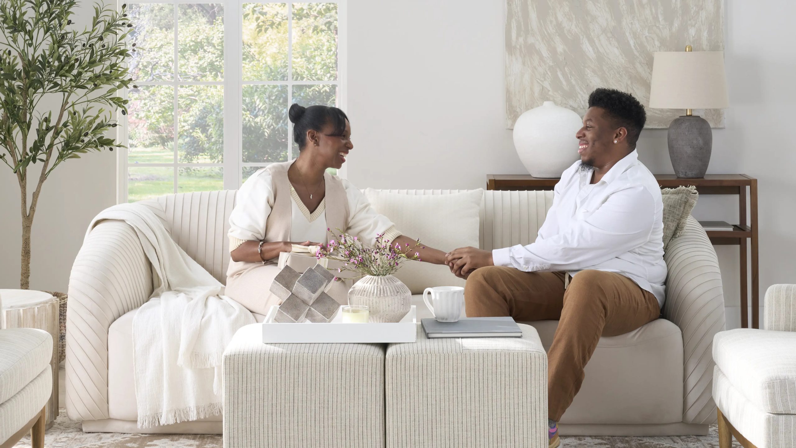 Woman and young man sitting together on a cream sofa sharing a gift in a bright neutral living room styled with Nourison home decor